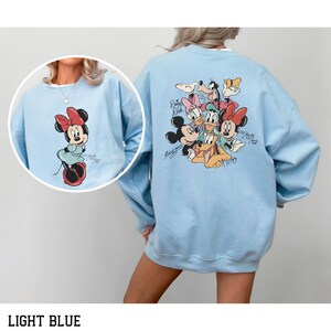 May include: Light blue sweatshirt featuring a Minnie Mouse graphic on the front and a group of Disney characters on the back. The Minnie Mouse graphic includes a red bow and dress. The back graphic includes Mickey Mouse, Donald Duck, and Pluto.