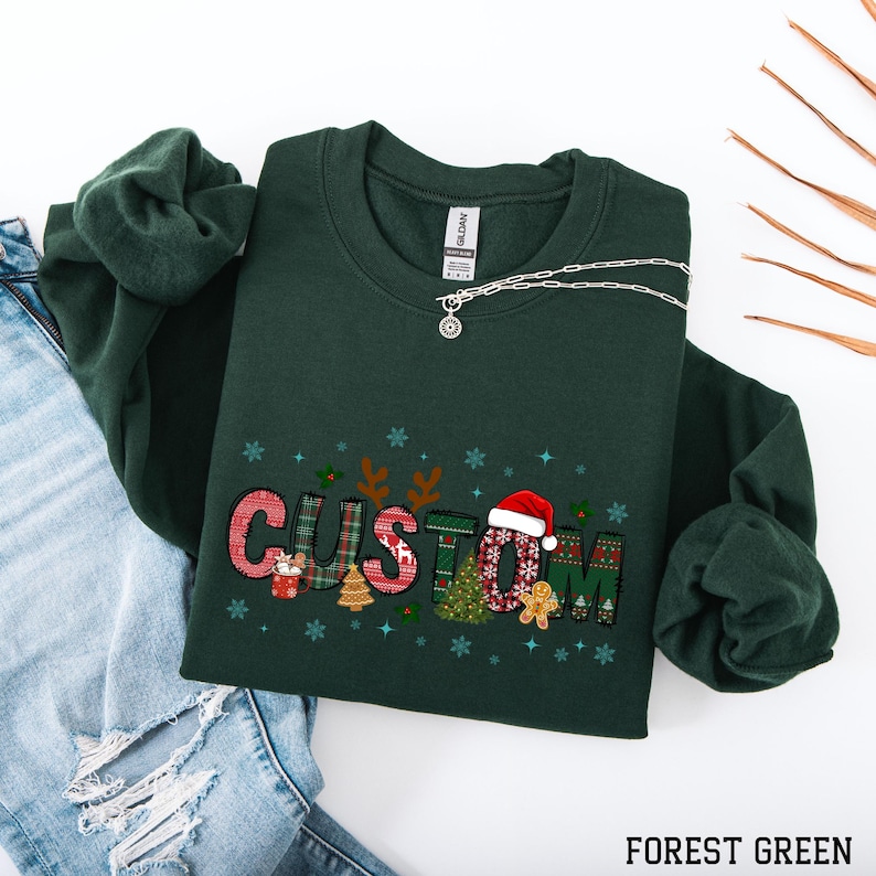 May include: Forest green sweatshirt with the word "CUSTOM" in a festive Christmas design. The letters are decorated with holiday patterns, a Santa hat, and gingerbread men. A silver necklace is draped across the sweatshirt.