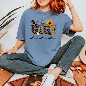 May include: A light blue t-shirt with a Halloween-themed design. The word "GIGI" is in the center, with a witch hat, bats, and a full moon. Below the word are the names "sarah", "riley", and "emma" with bat icons.