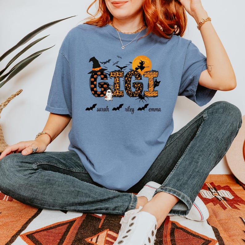 May include: A light blue t-shirt with a Halloween-themed design. The word "GIGI" is in the center, with a witch hat, bats, and a full moon. Below the word are the names "sarah", "riley", and "emma" with bat icons.