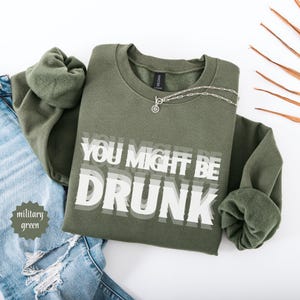 Puede incluir: Sudadera verde militar con el texto blanco "YOU MIGHT BE DRUNK". Un collar plateado con un colgante circular est&aacute; sobre la sudadera. La sudadera est&aacute; doblada sobre unos vaqueros azules.