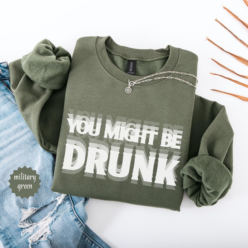 Puede incluir: Sudadera verde militar con el texto blanco "YOU MIGHT BE DRUNK". Un collar plateado con un colgante circular est&aacute; sobre la sudadera. La sudadera est&aacute; doblada sobre unos vaqueros azules.