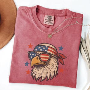 May include: A faded red short-sleeve t-shirt featuring a graphic of a bald eagle wearing a stars and stripes bandana and sunglasses. The shirt has a Comfort Colors brand label at the neck. Red and blue stars surround the eagle illustration.