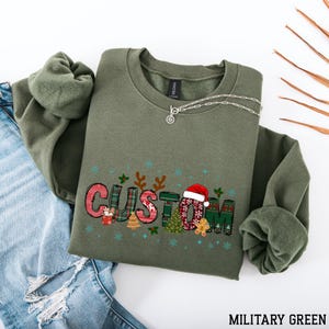 May include: An olive green sweatshirt with the word "CUSTOM" in a festive Christmas design. The design includes a Santa hat, gingerbread man, and Christmas tree. A silver necklace is draped across the sweatshirt. The text "MILITARY GREEN" is at the bottom.