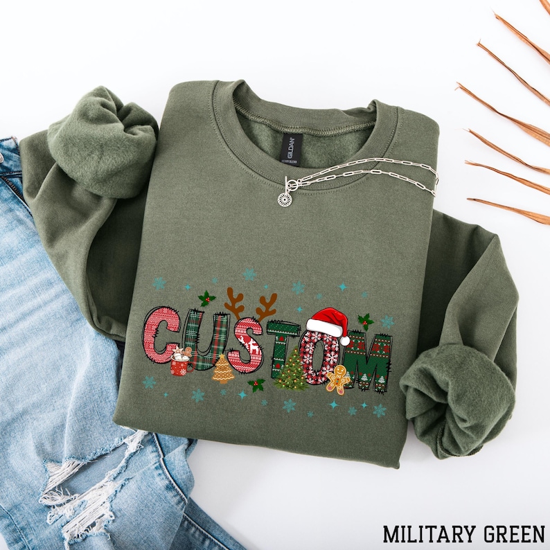May include: An olive green sweatshirt with the word "CUSTOM" in a festive Christmas design. The design includes a Santa hat, gingerbread man, and Christmas tree. A silver necklace is draped across the sweatshirt. The text "MILITARY GREEN" is at the bottom.