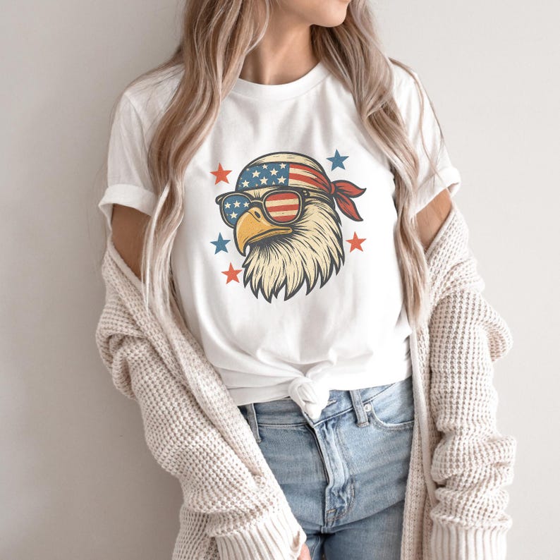 May include: A white short-sleeved t-shirt featuring a graphic of a bald eagle wearing a bandana and sunglasses with an American flag pattern. The eagle is surrounded by small red and blue stars. The shirt is tied at the waist and worn with jeans.