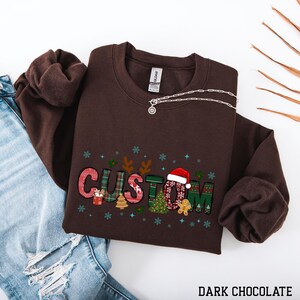 May include: A dark chocolate-colored sweatshirt with the word "CUSTOM" in a festive, patchwork design. The letters feature Christmas-themed patterns, including a Santa hat, gingerbread man, and Christmas trees. A silver necklace rests on the sweatshirt.