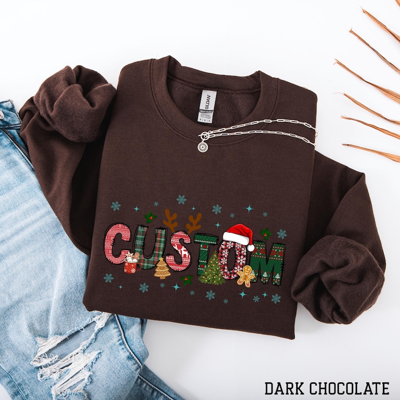 May include: A dark chocolate-colored sweatshirt with the word "CUSTOM" in a festive, patchwork design. The letters feature Christmas-themed patterns, including a Santa hat, gingerbread man, and Christmas trees. A silver necklace rests on the sweatshirt.