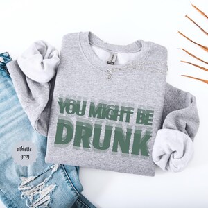 Puede incluir: Sudadera gris deportiva con el texto "YOU MIGHT BE DRUNK" en letras verdes superpuestas. La sudadera est&aacute; doblada, con las mangas dobladas para revelar un interior blanco. Una cadena plateada est&aacute; drapeada en la parte superior. Se ven unos vaqueros azules en el fondo.