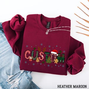 May include: A maroon sweatshirt with the word "CUSTOM" in a festive Christmas design. The letters are decorated with plaid, Christmas trees, and a Santa hat. A silver necklace is draped across the sweatshirt, which is laid on a pair of blue jeans.