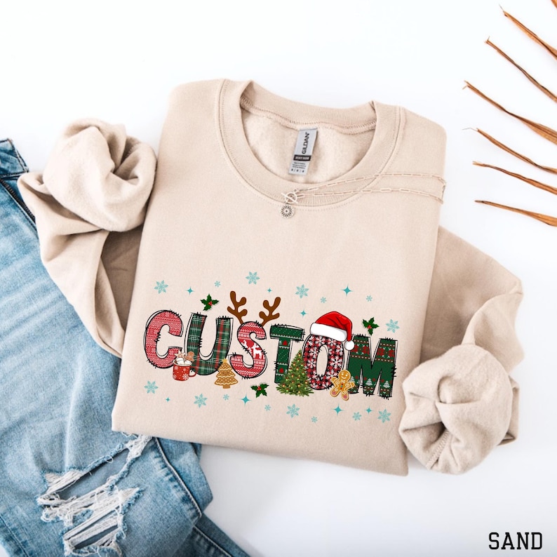 May include: Sand-colored sweatshirt with the word "CUSTOM" in a festive Christmas design. The letters are decorated with plaid patterns, a Santa hat, snowflakes, and gingerbread men. The sweatshirt is made of a soft material.
