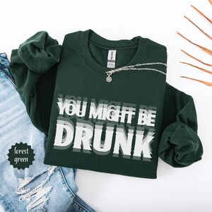 Puede incluir: Sudadera verde bosque con el texto "YOU MIGHT BE DRUNK" en blanco y gris. Un collar plateado con un colgante est&aacute; sobre la sudadera. La sudadera est&aacute; doblada sobre unos vaqueros azules.