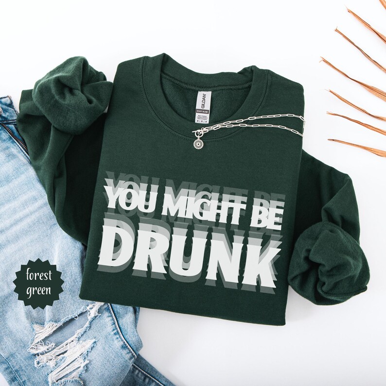Puede incluir: Sudadera verde bosque con el texto "YOU MIGHT BE DRUNK" en blanco y gris. Un collar plateado con un colgante est&aacute; sobre la sudadera. La sudadera est&aacute; doblada sobre unos vaqueros azules.