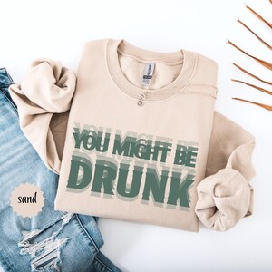 Puede incluir: Sudadera color arena con el texto "YOU MIGHT BE DRUNK" en letras verdes superpuestas. Se ve un collar plateado. La palabra "sand" est&aacute; en un peque&ntilde;o c&iacute;rculo a la izquierda. En el fondo se ven unos vaqueros azules.