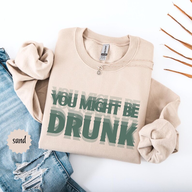 Puede incluir: Sudadera color arena con el texto "YOU MIGHT BE DRUNK" en letras verdes superpuestas. Se ve un collar plateado. La palabra "sand" est&aacute; en un peque&ntilde;o c&iacute;rculo a la izquierda. En el fondo se ven unos vaqueros azules.