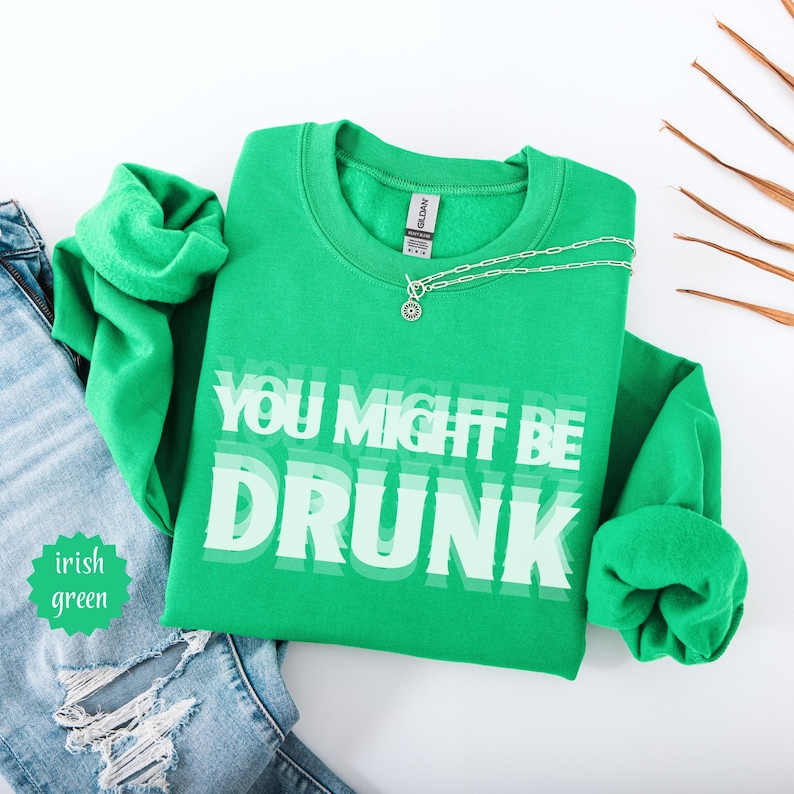 Puede incluir: Una sudadera verde irlandesa con el texto "YOU MIGHT BE DRUNK" en blanco. Un collar plateado con un colgante est&aacute; sobre la sudadera. La sudadera est&aacute; doblada junto a unos vaqueros azules.