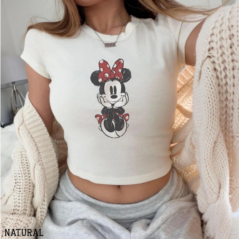 May include: Cream-colored, short-sleeved crop top featuring a Minnie Mouse graphic. The cartoon character is depicted in black and white with a red bow and accents. The word "NATURAL" is printed at the bottom of the image.