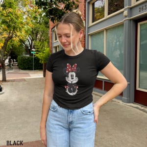 May include: Black t-shirt featuring a Minnie Mouse graphic with a red and white polka dot bow. The t-shirt is short-sleeved and has a crew neck. The word "BLACK" is printed at the bottom of the image.