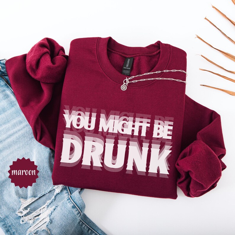 Puede incluir: Sudadera burdeos con cuello redondo con el texto "YOU MIGHT BE DRUNK" en blanco. Un collar plateado con un colgante est&aacute; drapeado sobre la sudadera. La sudadera se combina con unos vaqueros azules.