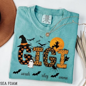May include: Sea foam green Comfort Colors t-shirt with a Halloween-themed design. The word "GIGI" is displayed with pumpkin and leopard print patterns. Includes bats, a witch on a broom, a ghost, and the names Sarah, Riley, and Emma.