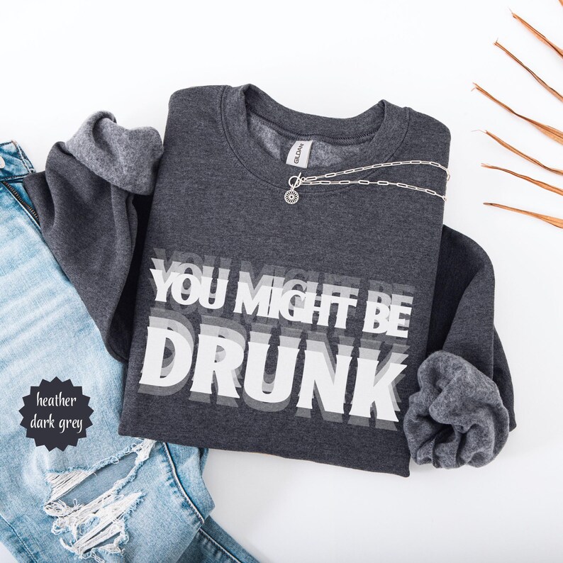 Puede incluir: Sudadera gris oscuro jaspeado con el texto "YOU MIGHT BE DRUNK" en una fuente blanca en capas. Un collar plateado con un colgante est&aacute; sobre la sudadera. La sudadera est&aacute; doblada junto a unos vaqueros azules.