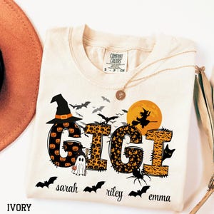 May include: Ivory-colored t-shirt with a Halloween-themed design. The word "GIGI" is displayed with pumpkin and leopard print patterns, a witch's hat, bats, and a witch silhouette. Additional names are printed below.