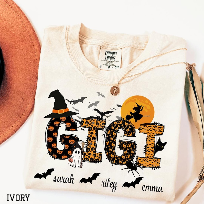 May include: Ivory-colored t-shirt with a Halloween-themed design. The word "GIGI" is displayed with pumpkin and leopard print patterns, a witch's hat, bats, and a witch silhouette. Additional names are printed below.