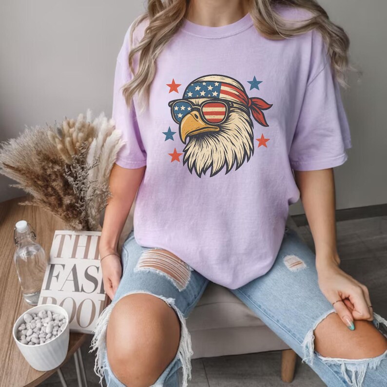 May include: A light purple short-sleeve t-shirt featuring a graphic of a bald eagle wearing American flag-patterned sunglasses and a matching bandana, surrounded by small red and blue stars. The shirt is paired with light blue distressed denim jeans.