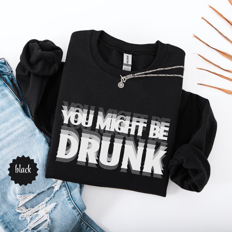 Puede incluir: Sudadera negra con la frase "YOU MIGHT BE DRUNK" en un dise&ntilde;o blanco y gris superpuesto. Un collar plateado con un colgante circular descansa sobre la sudadera. La palabra "black" est&aacute; en una estrella negra.