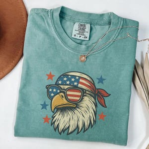 May include: A teal short-sleeved t-shirt featuring a graphic of a bald eagle wearing sunglasses and a bandana, both patterned with the American flag. The design includes red and blue stars. The shirt has a Comfort Colors label in size S.