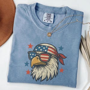 May include: A blue t-shirt featuring a graphic of a bald eagle wearing a stars and stripes bandana and sunglasses. The eagle is surrounded by red and blue stars. The shirt has a Comfort Colors label indicating size S, made in Honduras.