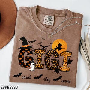 May include: Espresso-colored Comfort Colors t-shirt with a Halloween-themed design. The word "GIGI" is displayed with pumpkin and leopard print patterns, along with bats, a witch, and a ghost. The names "sarah", "riley", and "emma" are below.