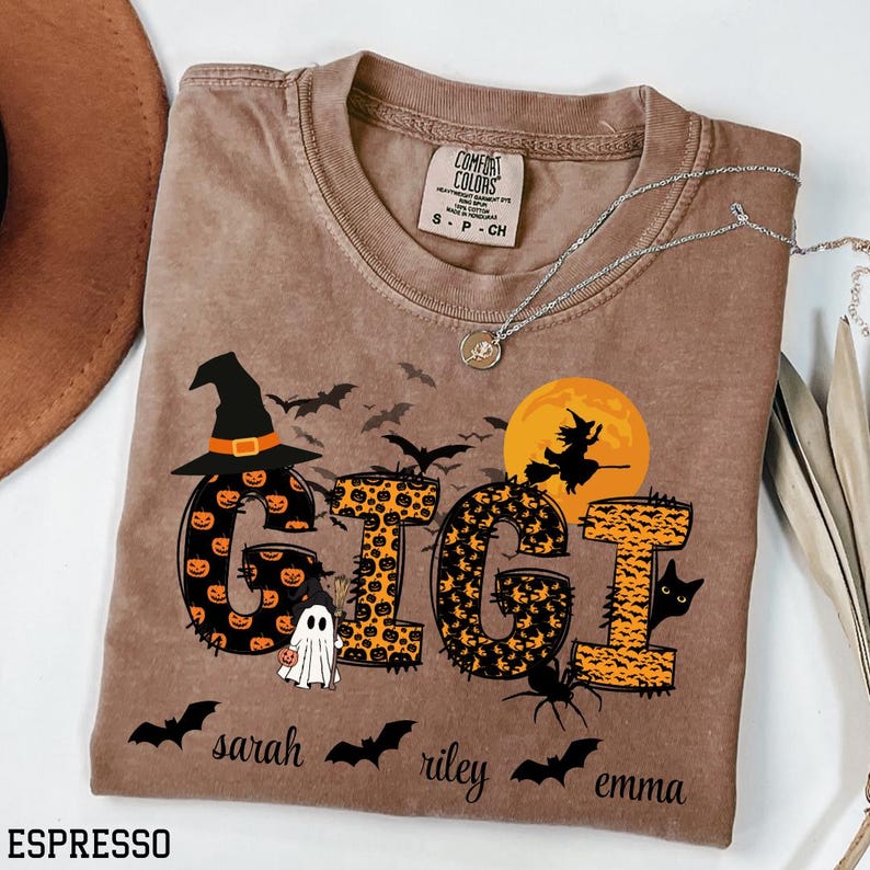 May include: Espresso-colored Comfort Colors t-shirt with a Halloween-themed design. The word "GIGI" is displayed with pumpkin and leopard print patterns, along with bats, a witch, and a ghost. The names "sarah", "riley", and "emma" are below.