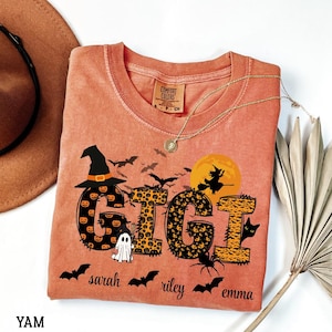 May include: Orange Comfort Colors t-shirt with a Halloween-themed design. The word "GIGI" is displayed with pumpkin and leopard print patterns, along with bats, a witch, a ghost, and the names "sarah", "riley", and "emma". A gold necklace is draped on the shirt.