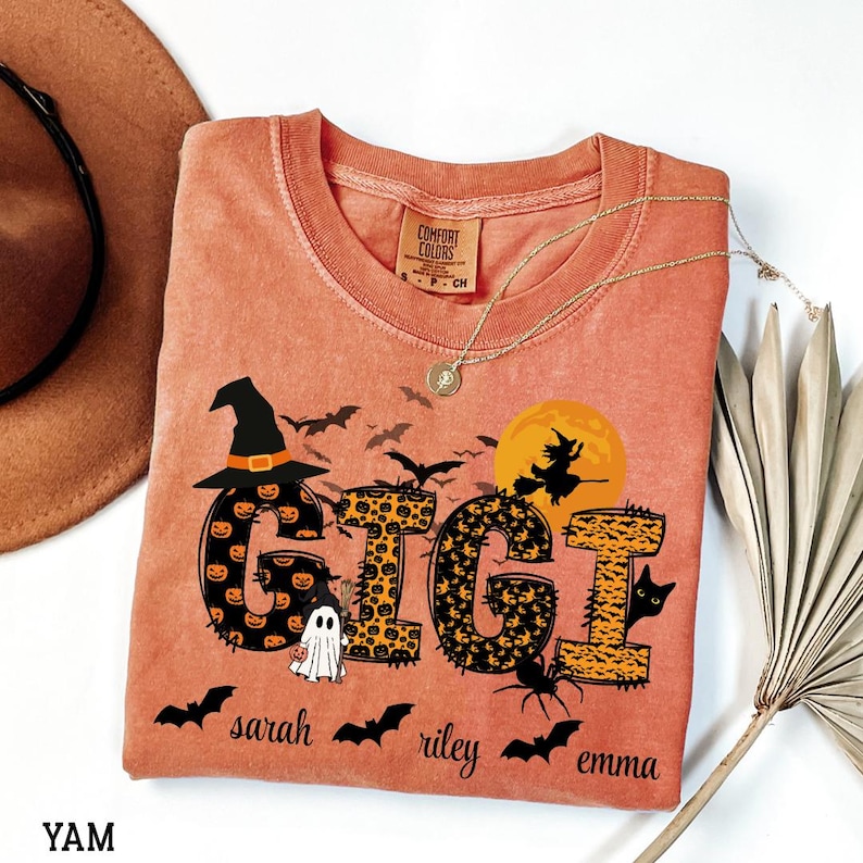 May include: Orange Comfort Colors t-shirt with a Halloween-themed design. The word "GIGI" is displayed with pumpkin and leopard print patterns, along with bats, a witch, a ghost, and the names "sarah", "riley", and "emma". A gold necklace is draped on the shirt.