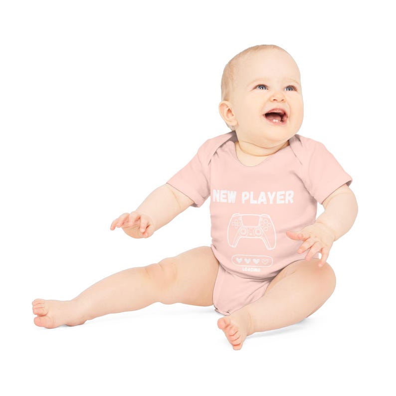 May include: A pink baby onesie with the text "NEW PLAYER" and a white outline of a video game controller. The onesie has a white outline of a heart with three white dots inside.