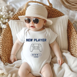 Könnte beinhalten: Ein Baby in einem weißen Body mit dem Text "NEW PLAYER" und einer Spielcontroller-Grafik. Das Baby trägt auch einen Strohhut und eine Sonnenbrille und liegt in einem Weidenkorb.
