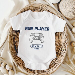 May include: White baby onesie with a blue graphic of a video game controller and the text "NEW PLAYER" and "LOADING" with a progress bar of hearts.