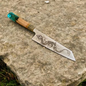 Könnte beinhalten: Ein handgefertigtes Kochmesser mit Holzgriff und einer mit einem Drachen gravierten Klinge. Der Griff hat ein türkisfarbenes Harzdetail und eine Messingzwinge. Das Messer wird auf einer strukturierten Steinoberfläche gezeigt.