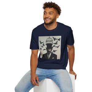 May include: Navy blue t-shirt featuring a surreal graphic of a man in a top hat with a birdcage on his head, surrounded by flying birds. The artwork is in black and white, creating a striking contrast against the shirt's color. The shirt is made of a soft material.