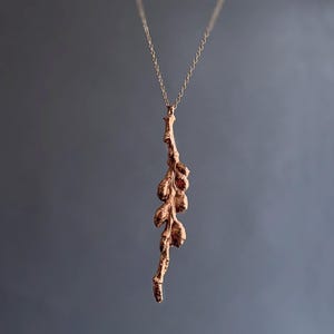 May include: A delicate, rose gold-colored necklace featuring a pendant shaped like a slender branch with textured, rounded buds. The pendant hangs from a fine chain, creating an elegant and naturalistic jewelry piece. The background is a muted gray.