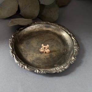 May include: A small, gold-toned flower brooch with detailed petals and a central accent, resting on a tarnished silver-colored decorative tray. The tray has an ornate, scalloped edge. Dried leaves are in the background.