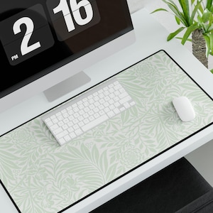May include: A white desk mat with a green floral pattern. The mat has a black border and is on a white desk. A computer keyboard, a computer mouse, and a clock are on the desk.