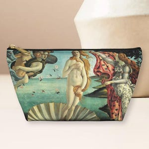 May include: A zippered makeup bag with a colorful print of a Renaissance painting depicting a woman standing on a seashell. The painting is titled "The Birth of Venus" by Sandro Botticelli.