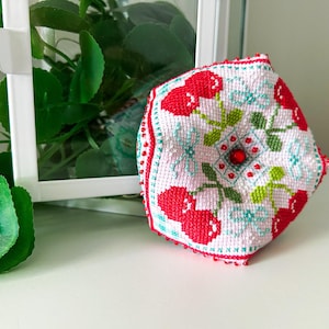 May include: A handcrafted, cross-stitched decorative item featuring a cherry design. The item is primarily white with red cherries, green leaves, and blue floral accents. A red bead is centered on the design, and the edges are trimmed with red beads.