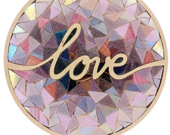 May include: Round mosaic art piece with the word "love" in a cursive font. The mosaic is made of small, irregularly shaped pieces in shades of pink, purple, and iridescent colors, set within a light wood frame.