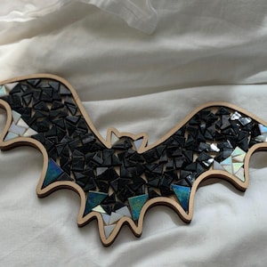 May include: A bat-shaped wall decoration featuring a wooden frame filled with a mosaic of small, triangular black tiles. Some tiles have an iridescent, reflective finish that displays hints of blue, green, and white light against the dark surface.