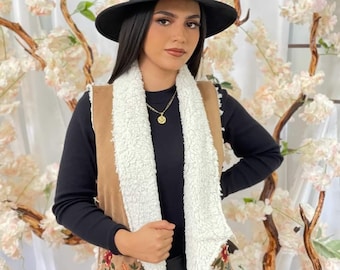 May include: A brown suede vest with a white faux fur collar and floral embroidery. The vest has a slightly asymmetrical hemline and is worn over a black long-sleeved shirt.