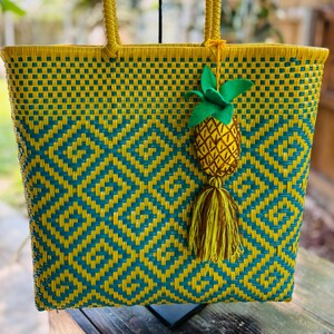 May include: A yellow and green woven tote bag with a pineapple charm. The bag has a geometric pattern and is made of plastic.