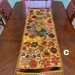 May include: A yellow table runner with embroidered floral designs in shades of red, brown, and white. The runner has a fringe border and is laid on a wooden table with three gold candle holders.